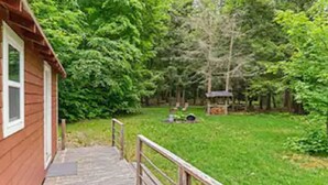 A wooden cabin with a wooden deck and a metal railing, surrounded by lush green trees and a grassy yard.