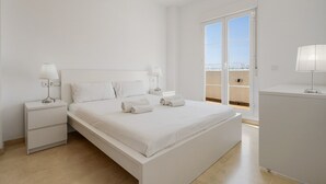 A minimalist white bedroom with a large bed, two bedside tables with lamps, and a view of the sky through a sliding glass door. - Nice Seaview 2 Bed 2 Bath Apartm. (Benalmádena)