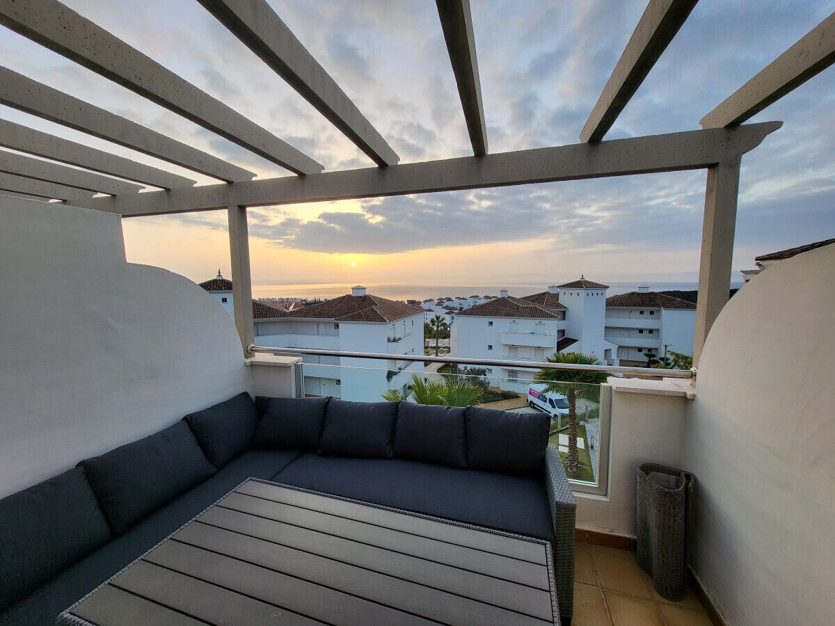 A balcony with a cushioned seating area, a view of buildings in the background, and a clear sky.