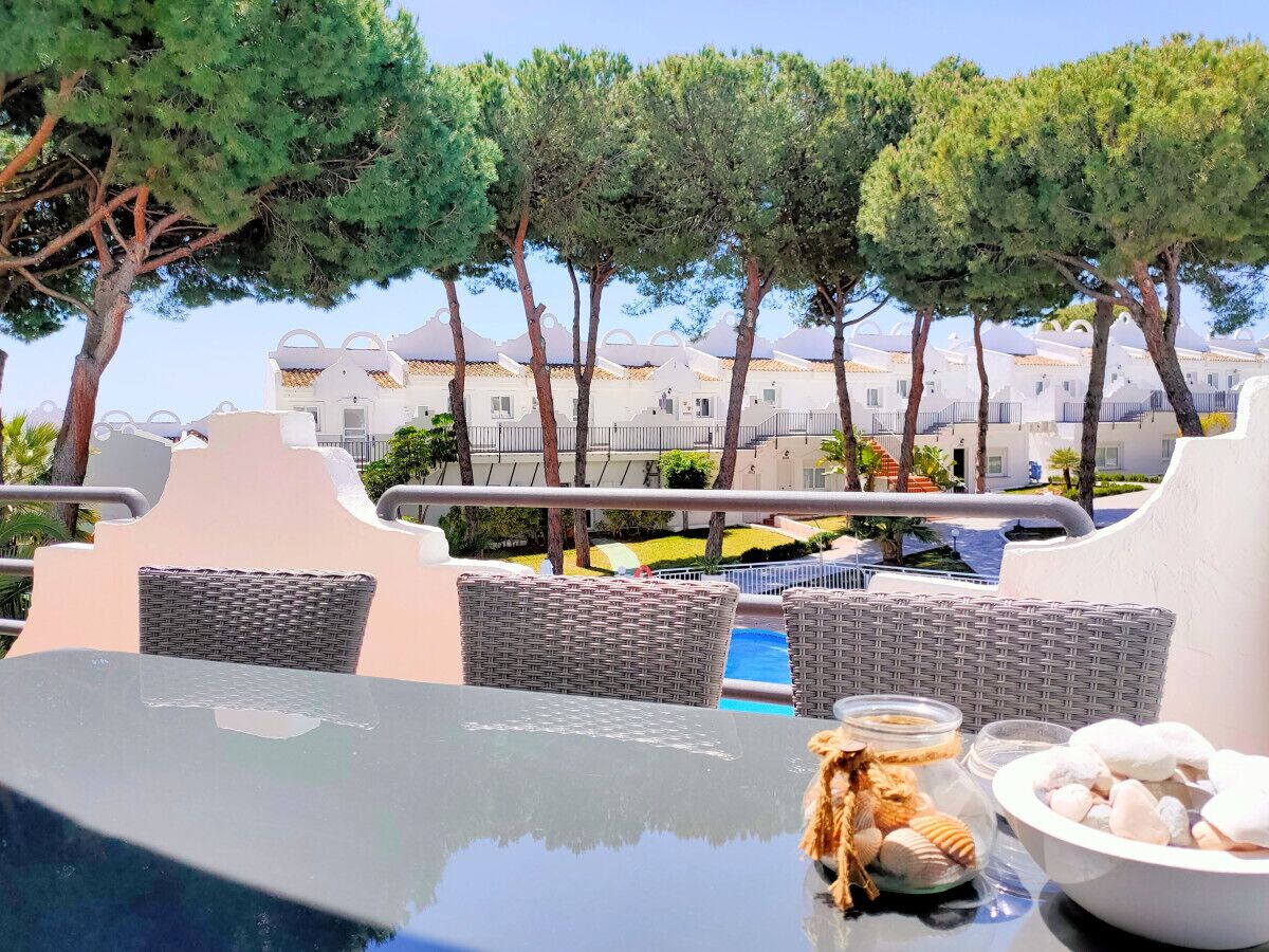 Un espace aménagé au bord de la piscine, avec des meubles en osier, une table en verre et une vue sur des bâtiments blancs en arrière-plan.