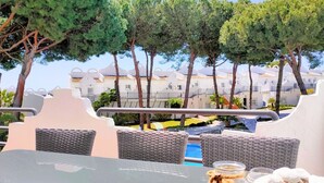 Zona junto a la piscina con mobiliario de mimbre, una mesa de vidrio y vistas a edificios blancos al fondo.