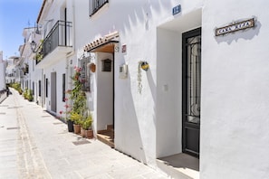A narrow street lined with white buildings, each with a black door and a small balcony. A sign reading 'CASA RIJA' is visible on one building.