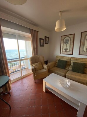 Una sala de estar con sofá, sillón y una mesa. Hay un balcón con vista al mar.