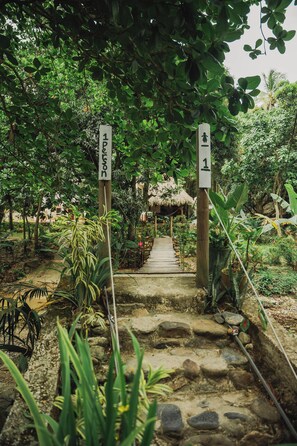 Un sendero serpentea a través de un exuberante jardín con escalones de piedra y postes de madera, rodeado de follaje denso y una estructura cubierta de paja.