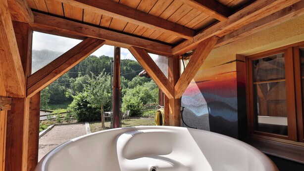 A wooden cabin with a large bathtub under a sloped roof, overlooking a forested area. - Chalet du Tanet in Soultzeren for 7 people spa sauna (Soultzeren)