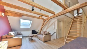 A loft-style living room with exposed wooden beams, a sofa set, a television, and a staircase. - Luxurious 500 m² villa with vaulted cellar, sauna, spa, large outdoor spaces (Sondernach)