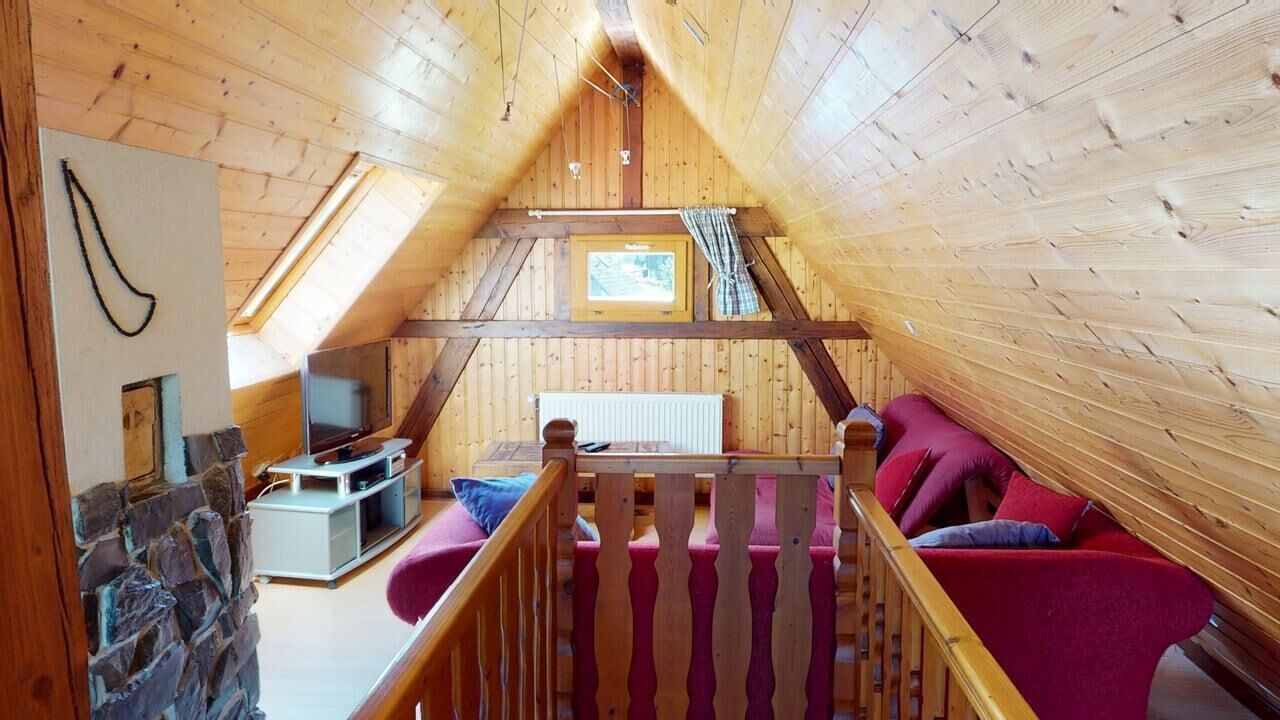 A cozy attic room with wooden walls and a sloped ceiling. It features a red sofa, a small TV, and a window with a view.