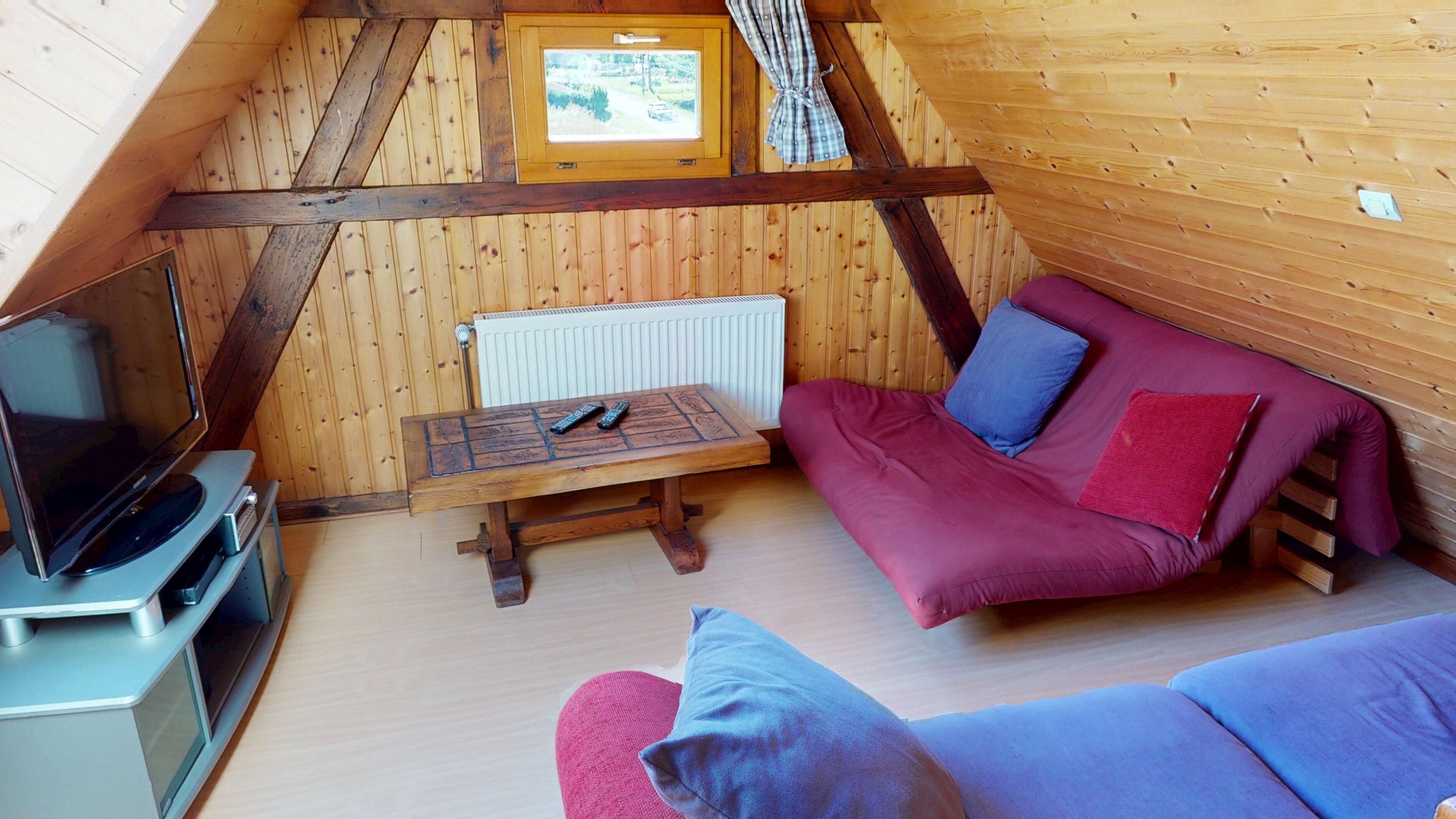 En mysig stuga med träpanel i taket, en soffa med kuddar, ett litet bord med en fjärrkontroll och en tv som står på ett stativ.