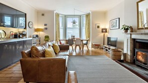Una sala de estar moderna con un sofá de cuero marrón, una chimenea, un comedor con mesa y sillas, y una cocina con un ventanal grande y un espejo.
