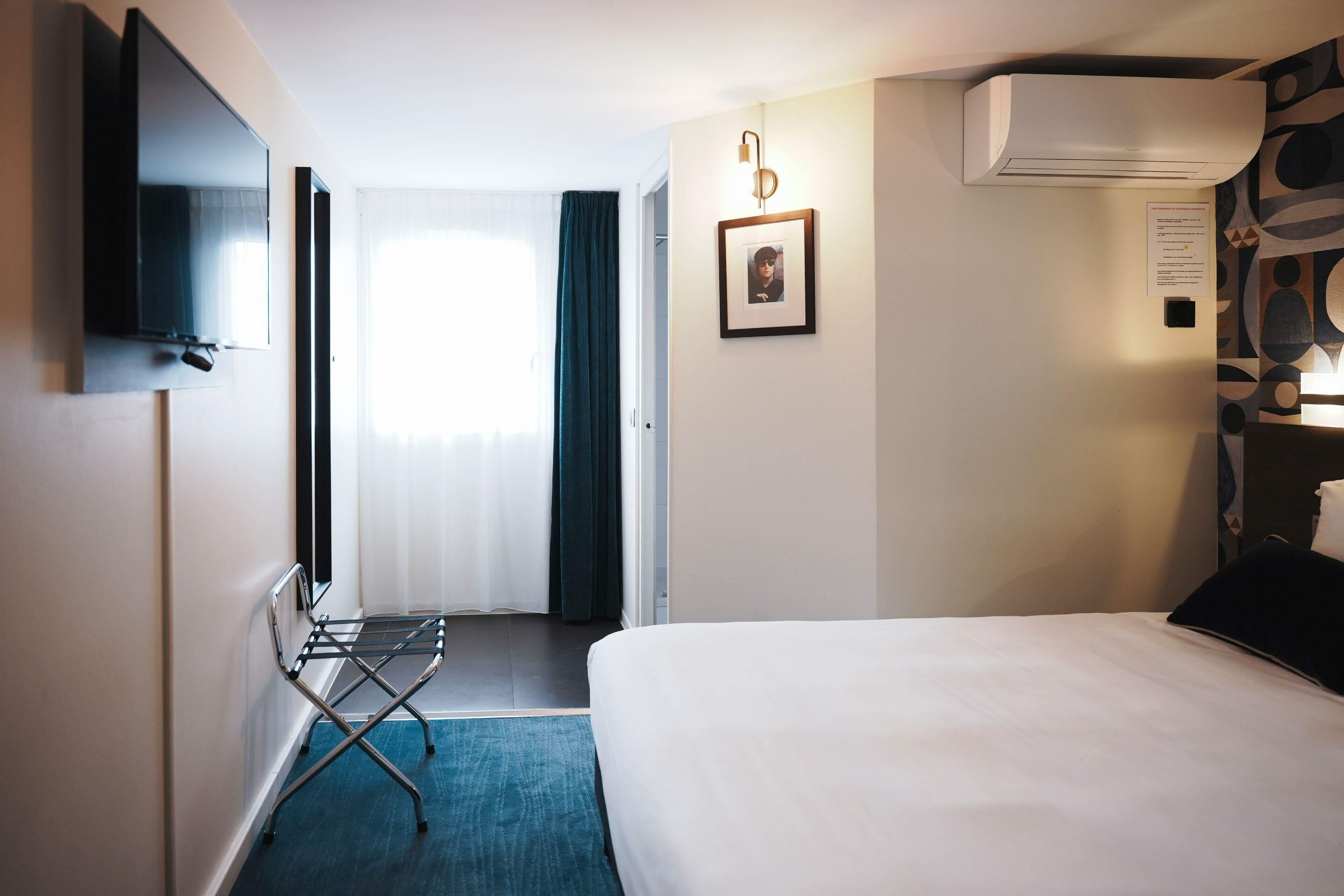 Une chambre d’hôtel avec un lit, un téléviseur fixé au mur, un appareil de climatisation, un cadre photo et une fenêtre avec des rideaux.