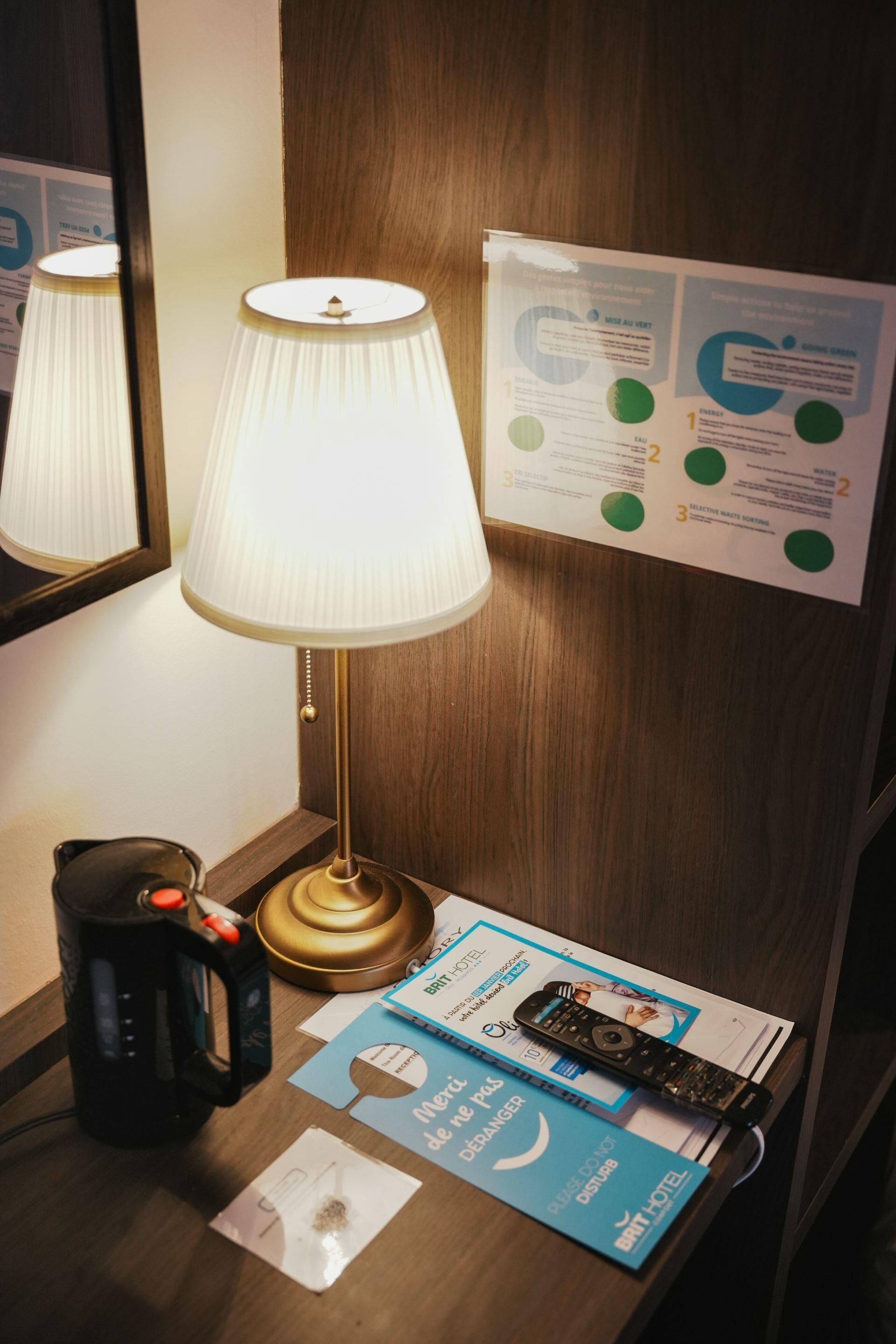 A hotel room with a wooden desk, a lamp, a kettle, a remote control, and a printed guide on the wall.