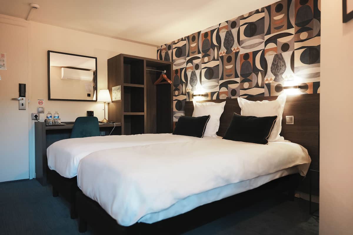A hotel room with a large bed, a desk with a chair, a wardrobe, and a wall with abstract art.