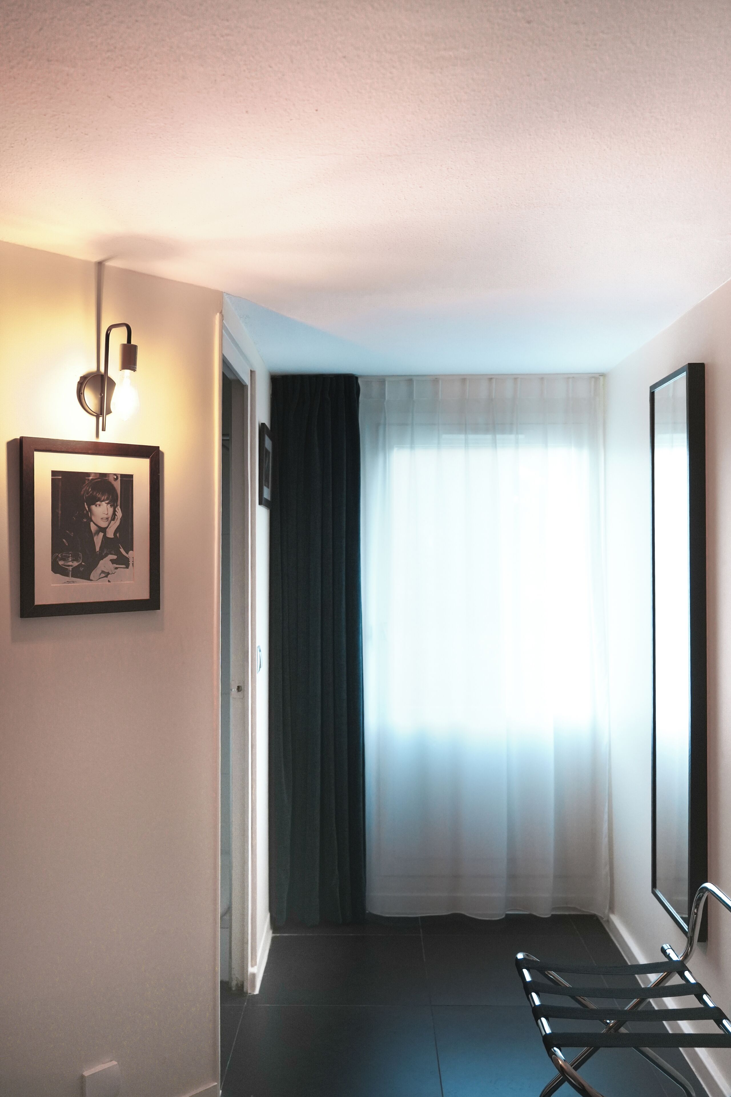 A hallway with a framed picture on the wall, a light fixture, a window with sheer curtains, and a metal chair.