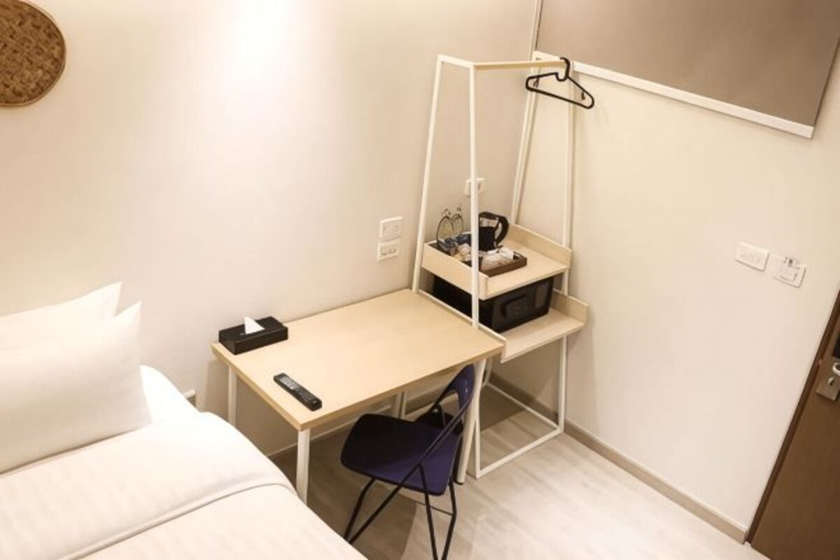 A compact hotel room with a bed, a desk, a chair, a hanging coat rack, and a small shelf with items.