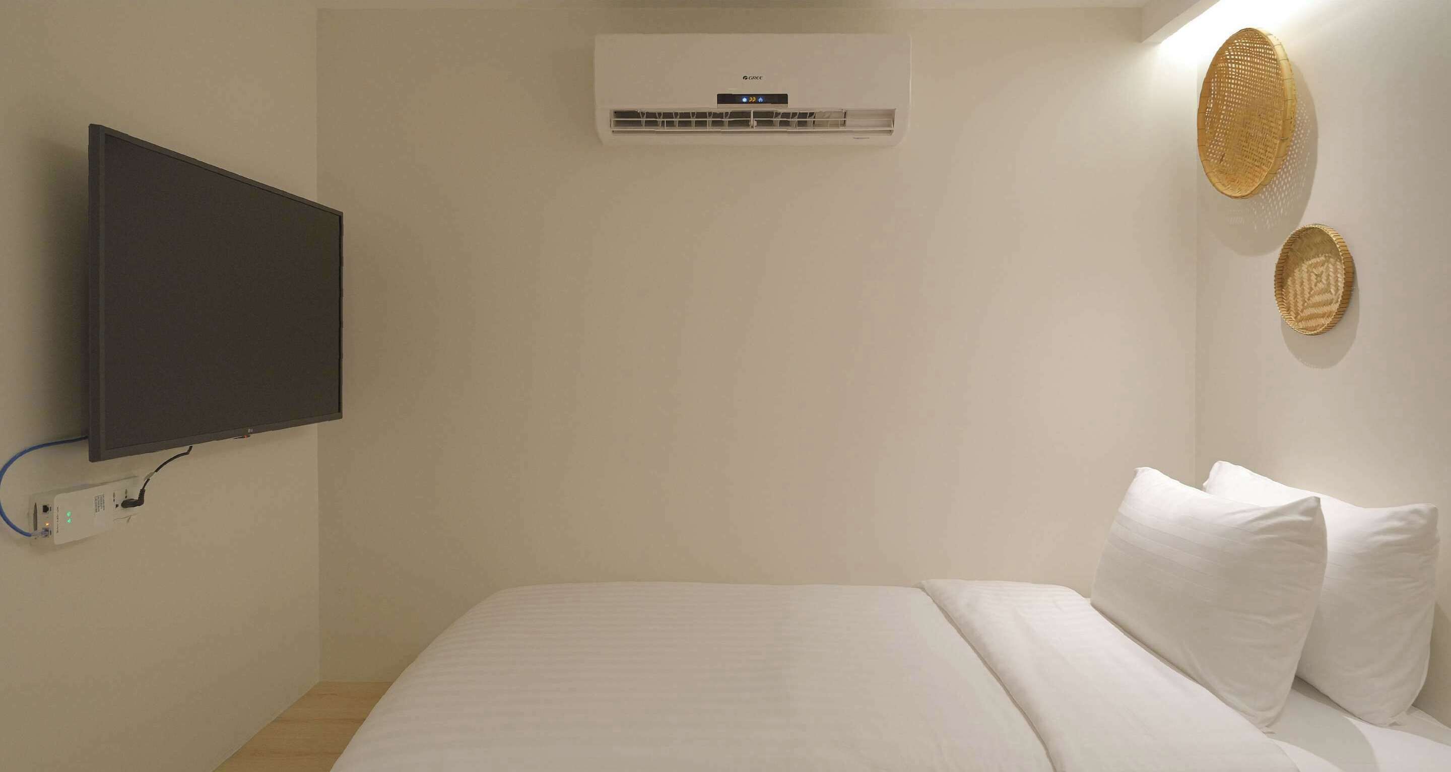 A bedroom with a bed, a wall-mounted air conditioner, a flat-screen TV, and two woven wall decorations.