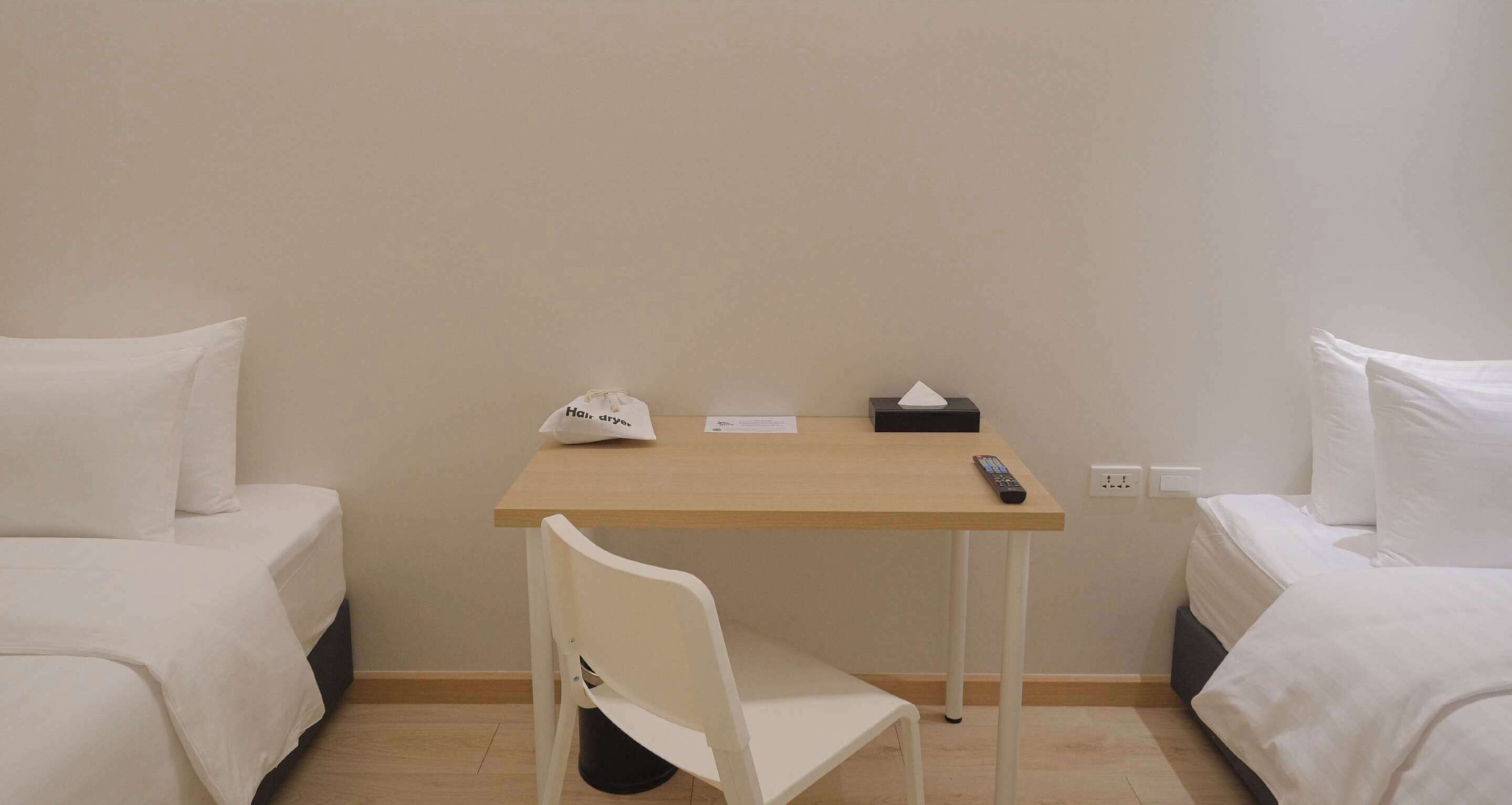 Ein kleines Zimmer mit zwei Betten, einem Schreibtisch, einem Föhn, einer Fernbedienung und einem Papierkorb.
