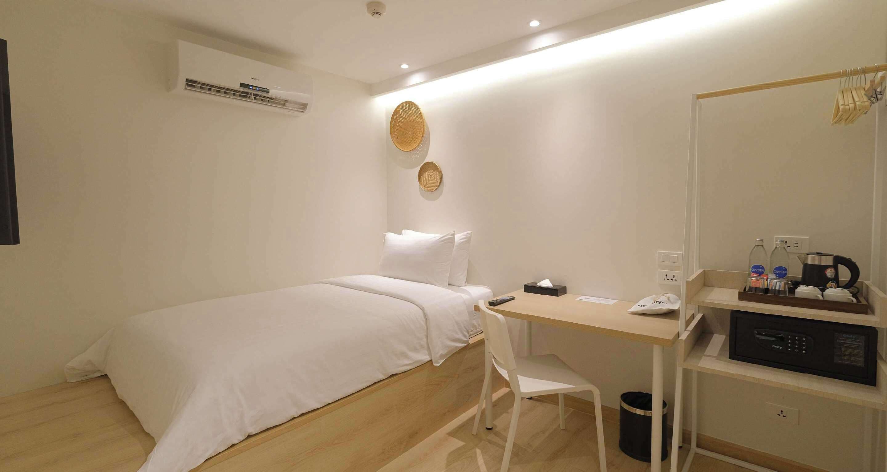 A hotel room with a bed, a desk, a chair, a trash bin, a microwave, and a shelf with toiletries.