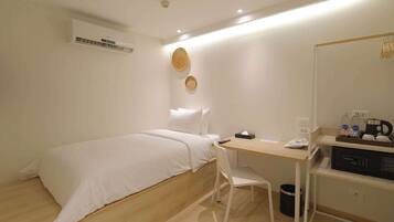 A hotel room with a bed, a desk, a chair, a trash bin, a microwave, and a shelf with toiletries.