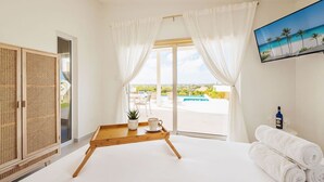 Et moderne hotelværelse med en stor seng, et træsidebord med en plante og en kop, et fjernsyn på væggen og udsigt til en pool og grøn bevoksning udenfor.