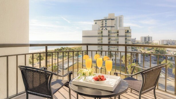A balcony with two wicker chairs, a small table with a tray of food and drinks, and a view of a beach and cityscape. - New Listing! Meridian Plaza Resort #606 (Myrtle Beach)