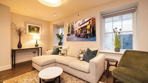 A living room with a sectional sofa, a small round coffee table, a green armchair, a painting of a street scene, and a potted plant by the window.