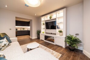 Una sala moderna con un sofá, una mesa de centro y una chimenea. Se observa una cocina al fondo con un lavamanos y armarios.