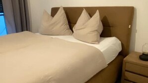 A neatly made bed with beige bedding and pillows, a wooden nightstand with drawers, and a window with curtains.