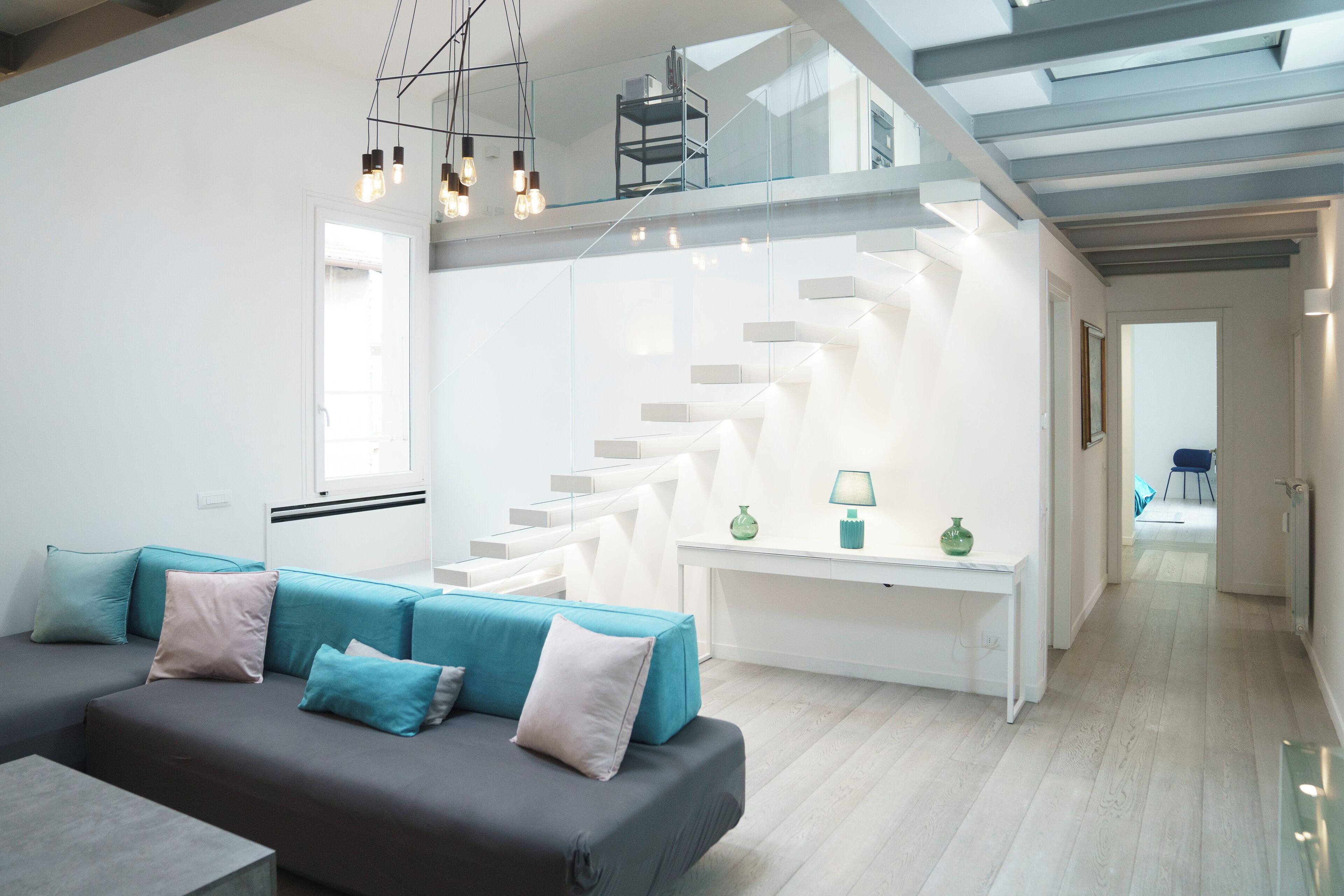 A modern living room with a grey sofa, teal cushions, a white console table, and a staircase leading to an upper level.