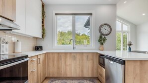 A modern kitchen with wooden cabinets, a built-in dishwasher, a large window with a view of trees, and a clock on the wall.