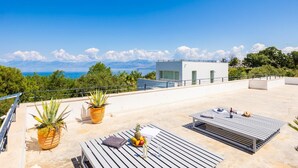 Eine Dachterrasse mit Außenbestuhlung, Blick auf das Meer und ein modernes Gebäude im Hintergrund.