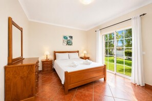 A bedroom with a wooden bed, two bedside tables with lamps, a wooden dresser, a large window with a view of greenery, and a ceiling light.