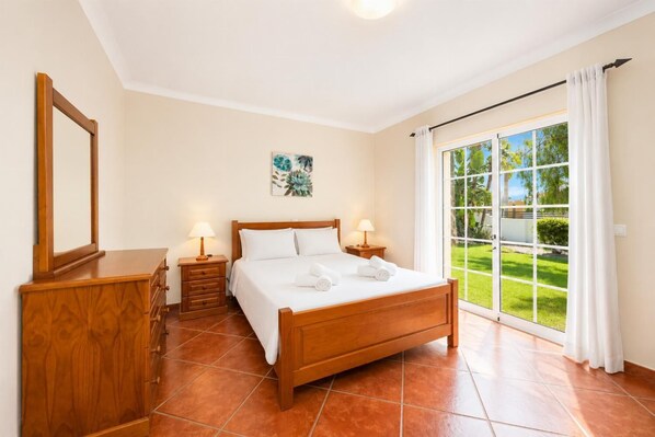 A bedroom with a wooden bed, two bedside tables with lamps, a wooden dresser, a large window with a view of greenery, and a ceiling light. - Casa Amendoeira Large Private Pool, WiFi (Albufeira)