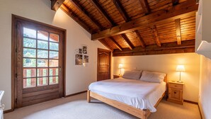 Un dormitorio con una cama de madera, dos mesitas de noche con lámparas, una puerta de madera y un ventanal con vistas a zonas verdes.