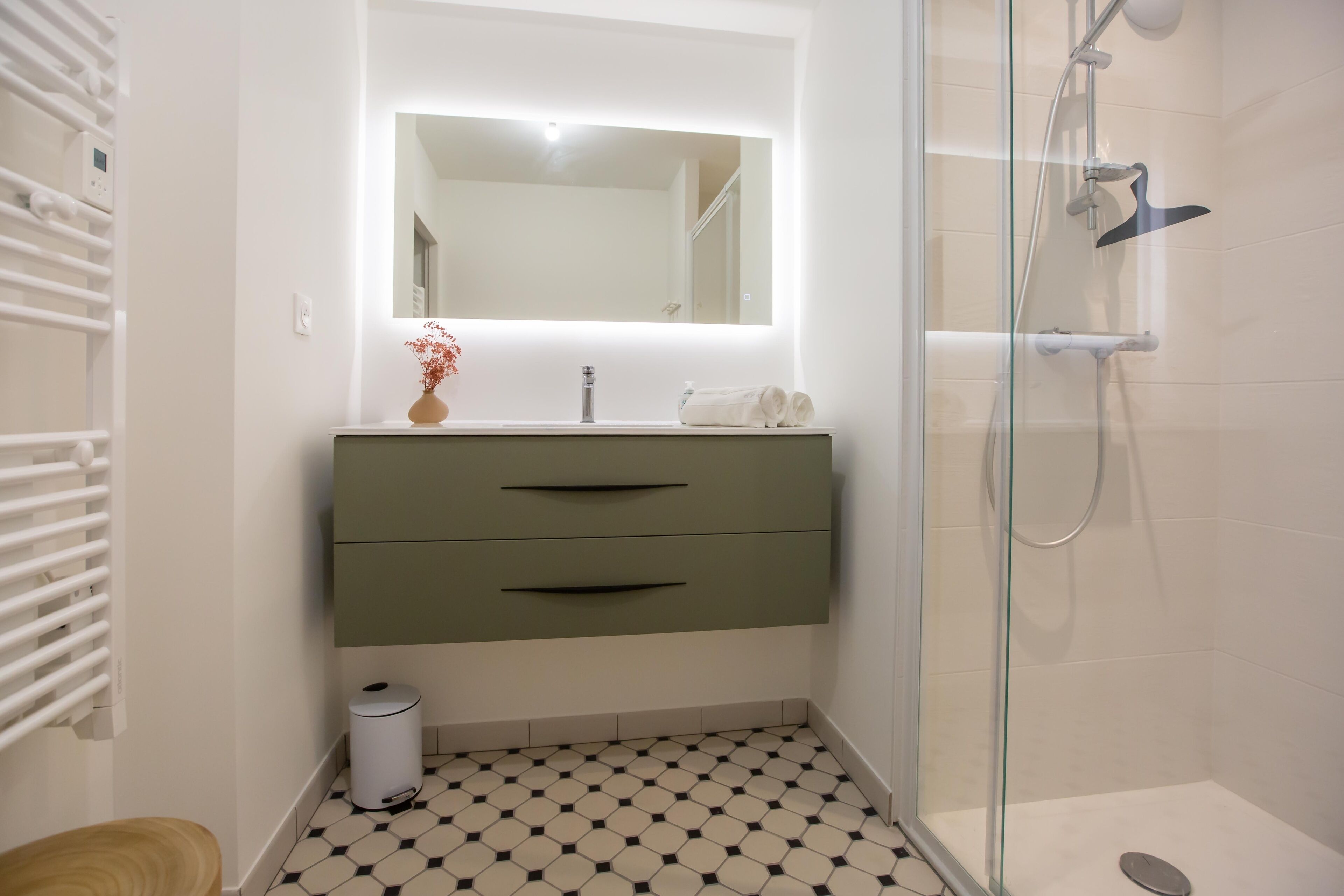 Une salle de bain au design moderne, dotée d’une douche à parois de verre, d’un lavabo blanc avec un miroir au-dessus, et d’un carrelage au motif original.