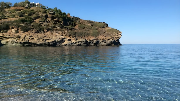 Un paisaje costero con aguas cristalinas, terreno rocoso y una colina de vegetación escasa.