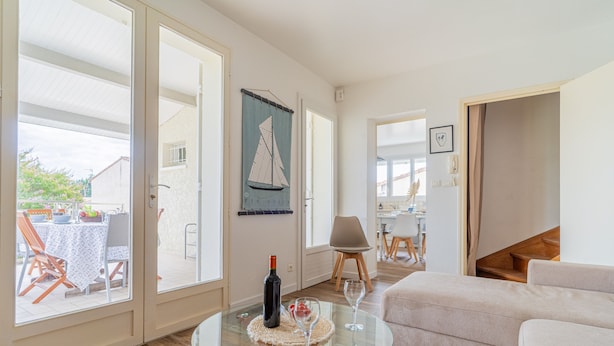 A spacious living area with a glass coffee table, a sofa, and a dining table with chairs. There is a sailboat decoration on the wall and a bottle of wine on the table. - Ecrin Marin - apt terrasse, bassin d'arcachon (La Teste-de-Buch)