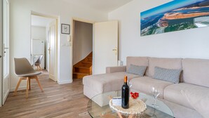A living room with a beige sofa, a glass coffee table, a bottle of wine, and a painting of a landscape. - Ecrin Marin - apt terrasse, bassin d'arcachon (La Teste-de-Buch)
