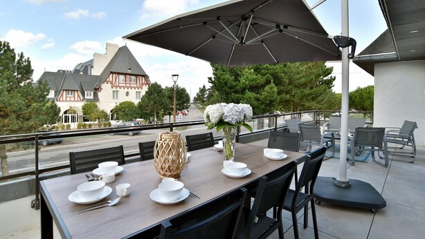 Un área de comedor al aire libre con una mesa dispuesta para cuatro, un paraguas grande y vista a un edificio tradicional en el fondo.