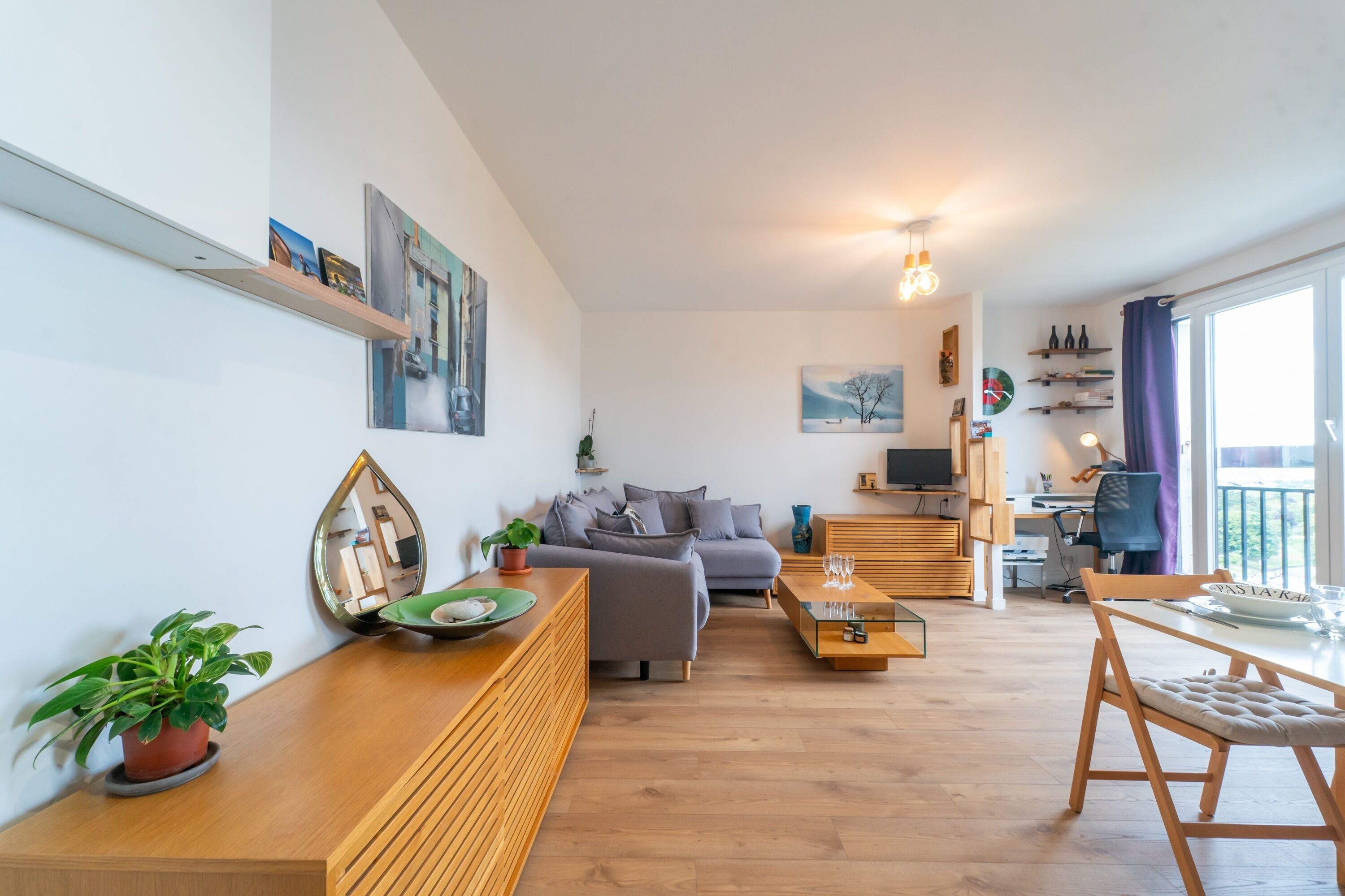 A modern living room with a grey sofa, wooden coffee table, and a dining area with a table and chairs. There are wall-mounted shelves with decorative items and a large window with curtains.