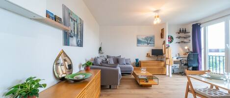 A modern living room with a grey sofa, wooden coffee table, and a dining area with a table and chairs. There are wall-mounted shelves with decorative items and a large window with curtains.
