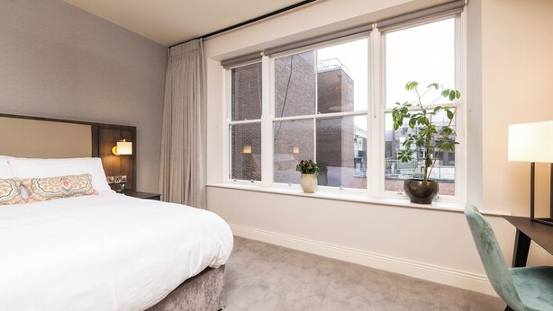 A hotel room with a large bed, a window with a view of buildings, a small table, and a chair. - Spacious 1 Bed Loft Apartment in Dublin City Centre (Dublin)