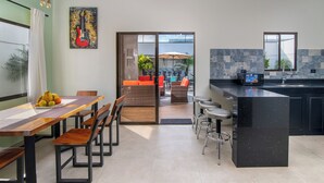 A modern kitchen with a black countertop, a dining area with wooden chairs, and a view of the outdoors through a glass door. - La Casa de Papi (Parrita)