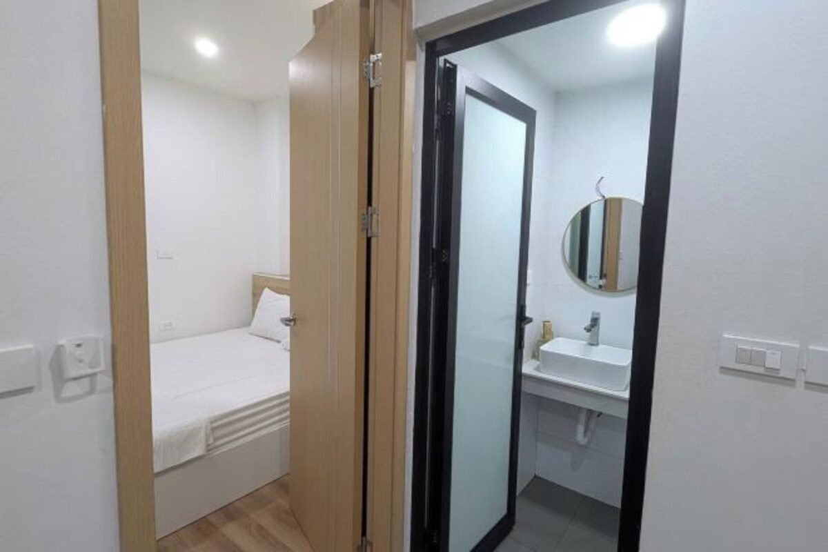 A small, clean room with a bed, a bathroom with a sink and mirror, and a door leading to another room.