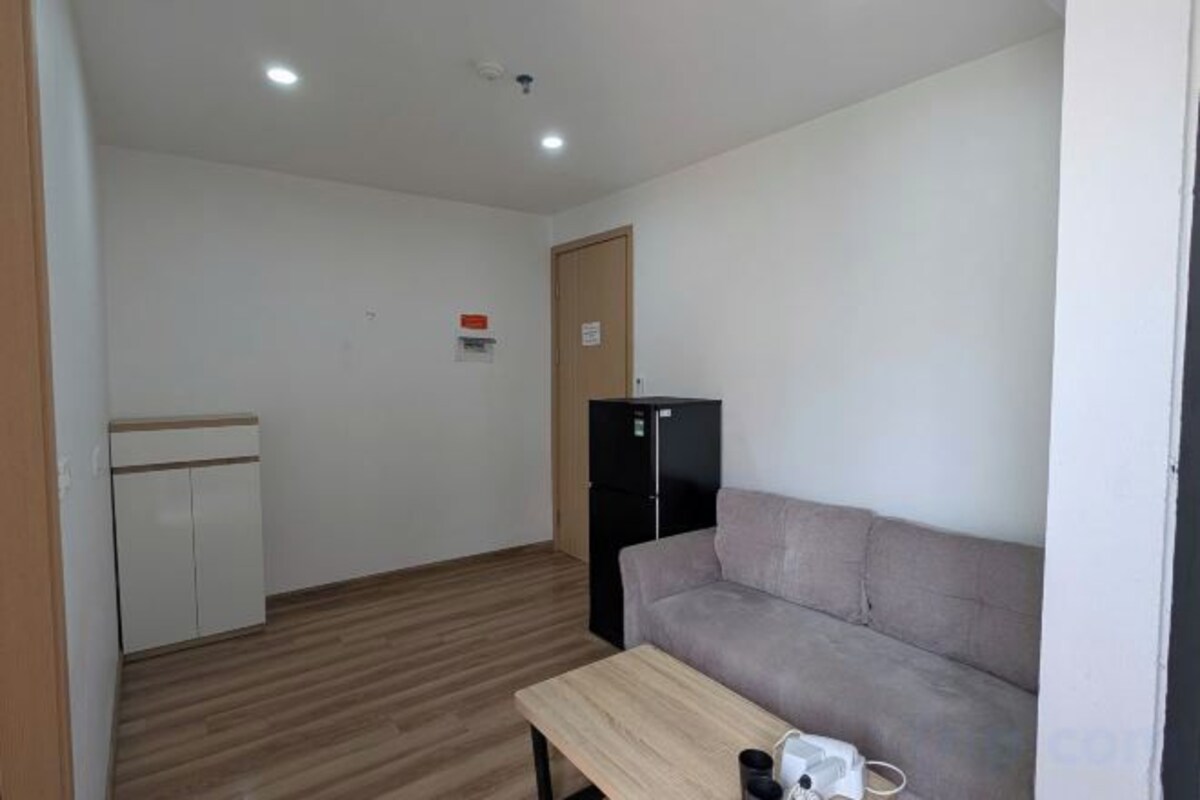 Una habitación con piso de madera, un sofá, una mesita, un refrigerador y una zona de kitchenette.