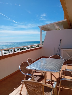 Un balcon avec une table en métal et des chaises en osier, donnant sur une plage bordée de palmiers et sur l’océan.