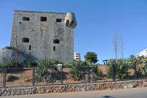 Una torre de piedra con múltiples ventanas y base de piedra, rodeada por una valla y diversas plantas.