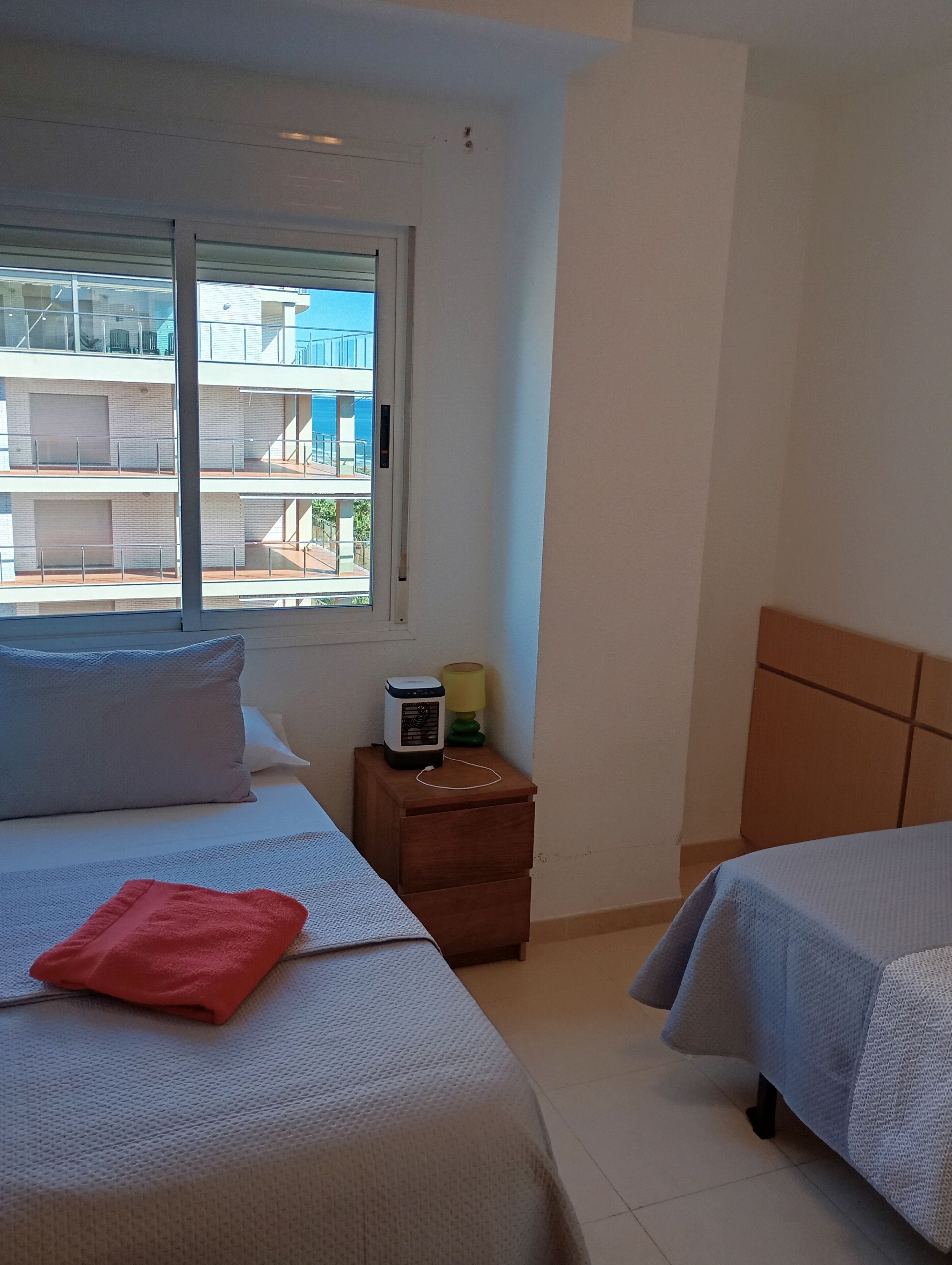 Een hotelkamer met twee bedden, een nachtkastje met een lamp en een raam met uitzicht op een gebouw.