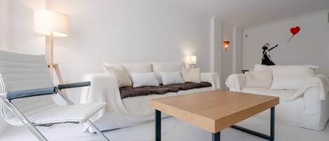 A modern living room with a white sofa, a wooden coffee table, a white armchair, and a wall with a red heart decoration.