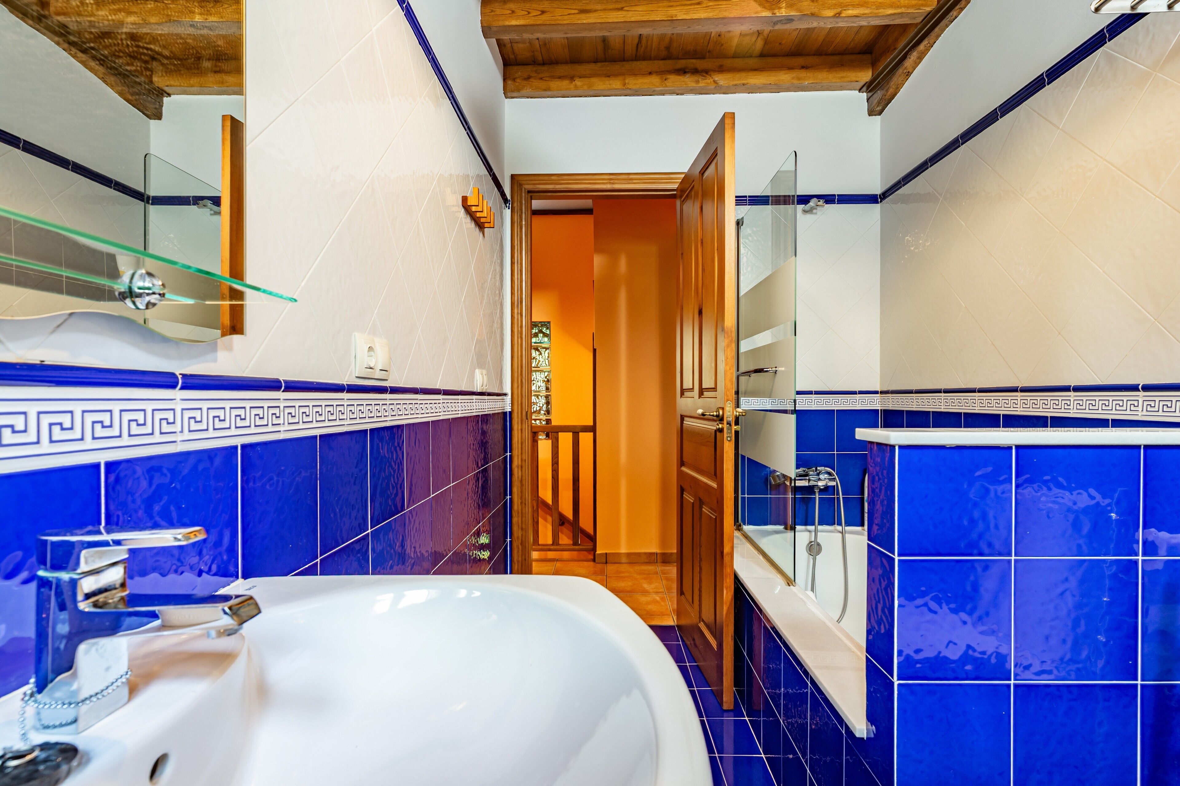 Ein Badezimmer mit einem weißen Waschbecken, blauen Fliesen, einer Glasablage und einer Holztür zu einem anderen Raum.