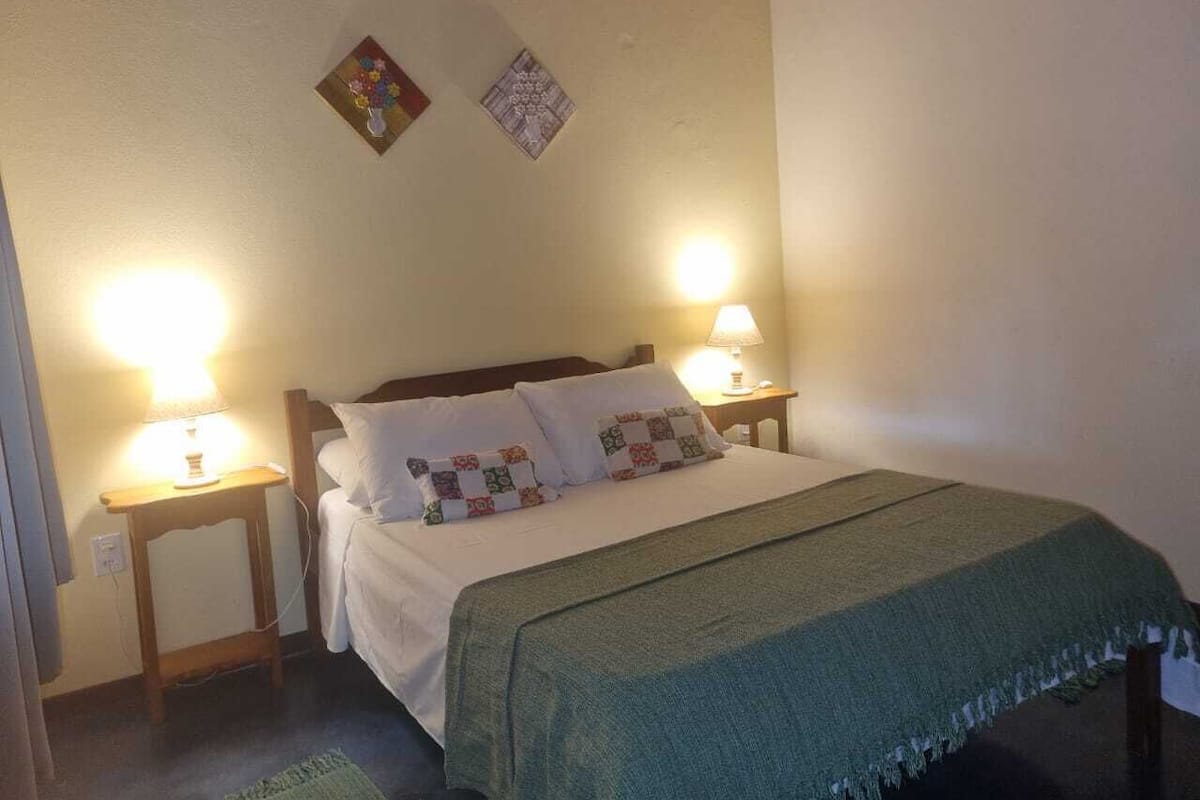 A neatly made bed with a green blanket, two bedside lamps, and two wall-mounted pictures.